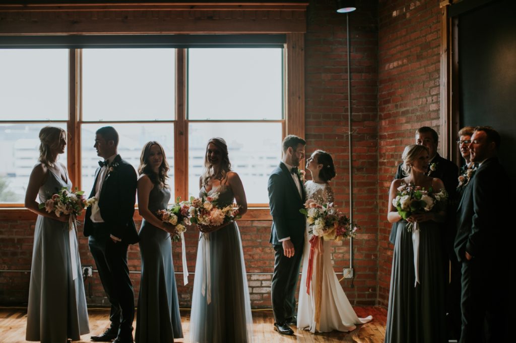 a window in a brick wall at the mavris backlights a bride and groom and their bridal party as they stand posed