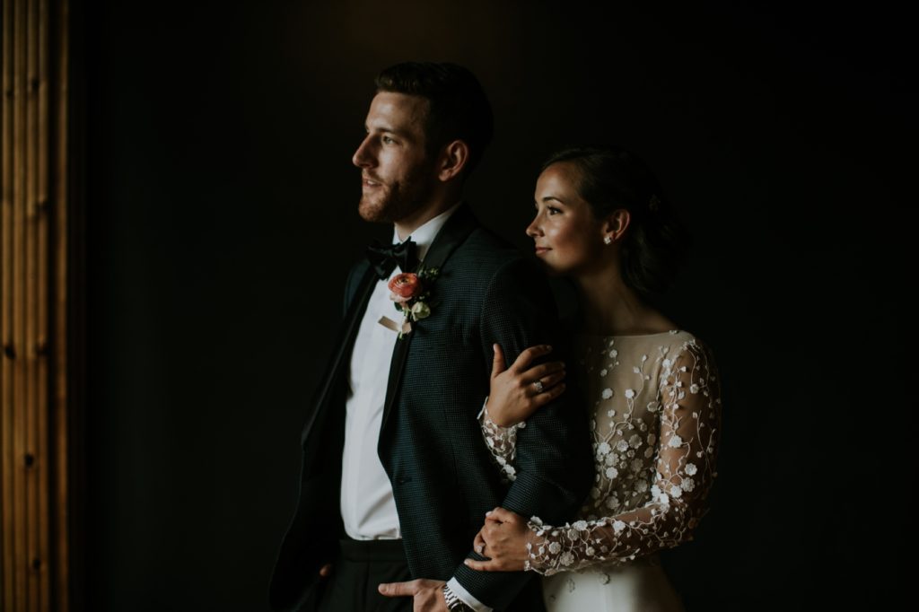 bride in modern chic wedding dress stands with groom in two piece tux in moody window light, woman behind man and clutching his arm