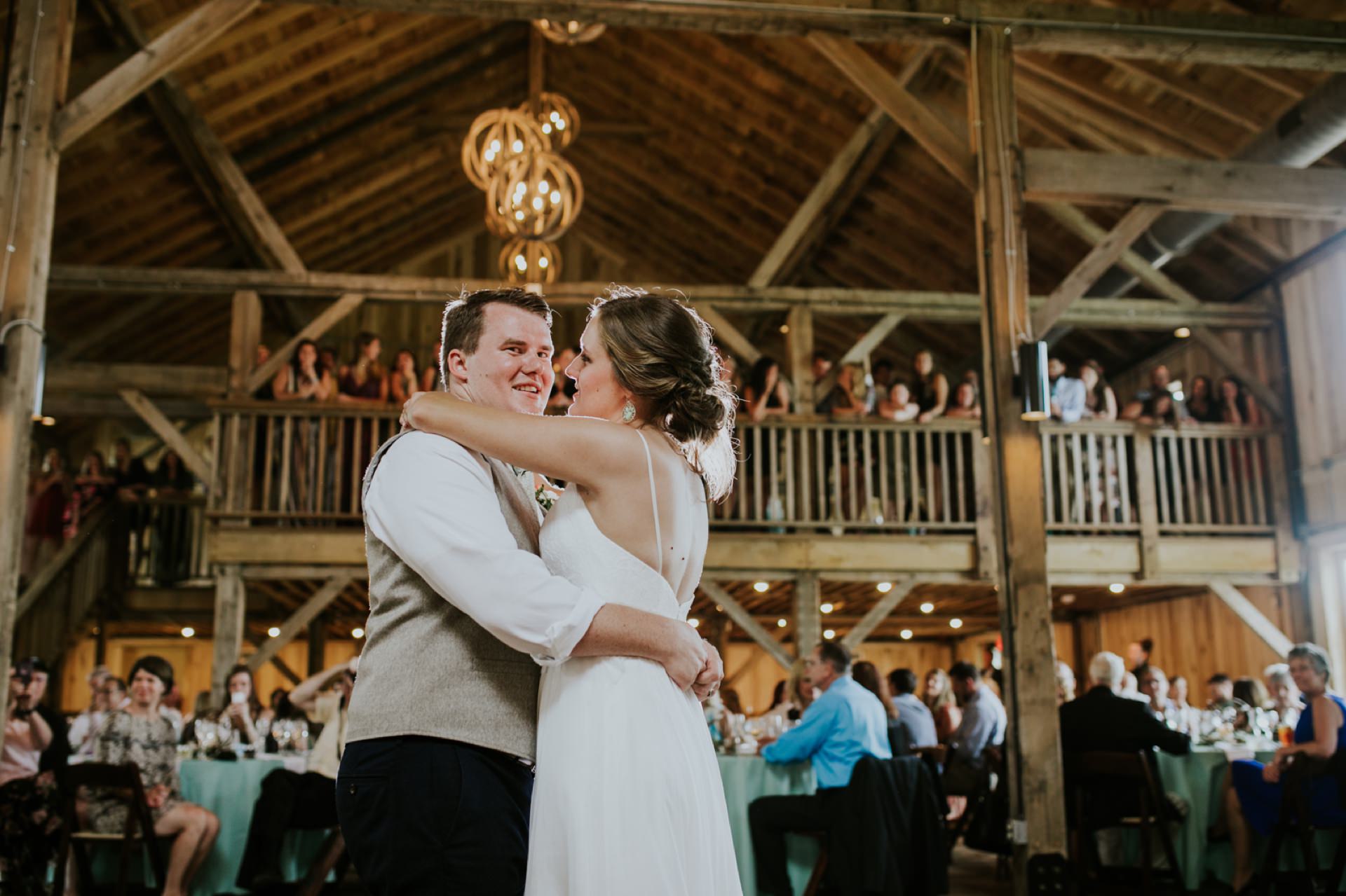 a bride and groom dance in a barn at their lindley farmstead wedding while people watch on the ground floor and in the balcony