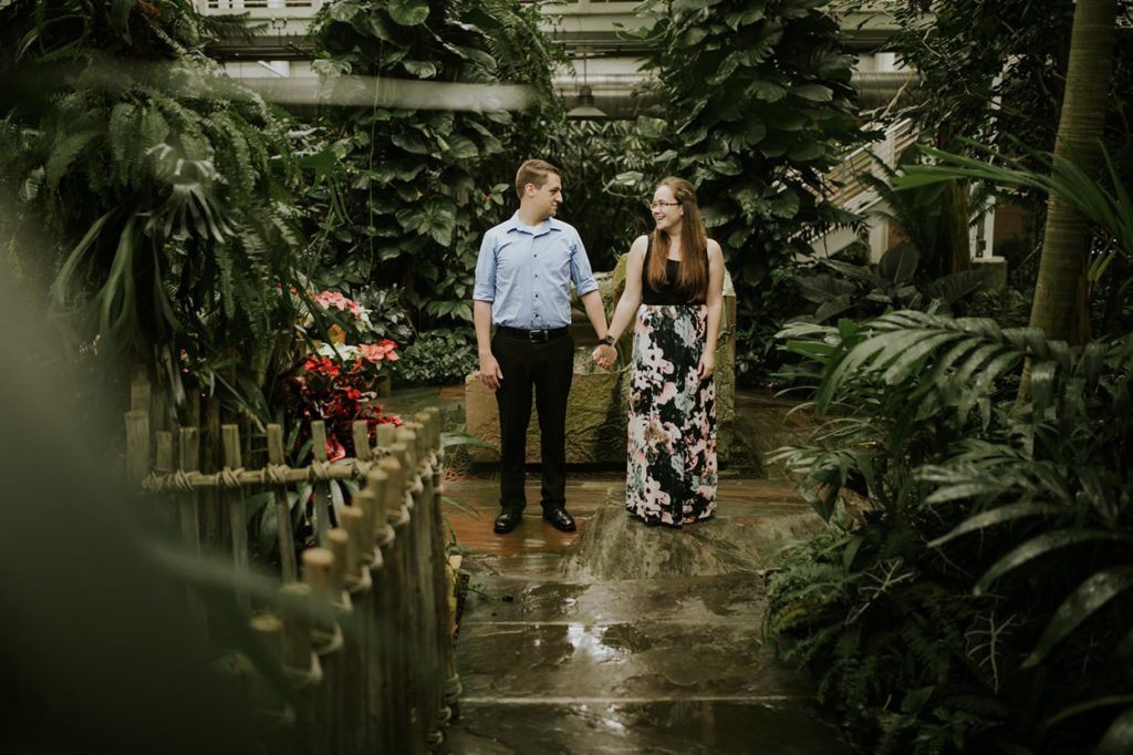 Man in slacks and dress shirt holds hands with woman in sun dress at their botanical gardens engagement session at the Indianapolis zoo