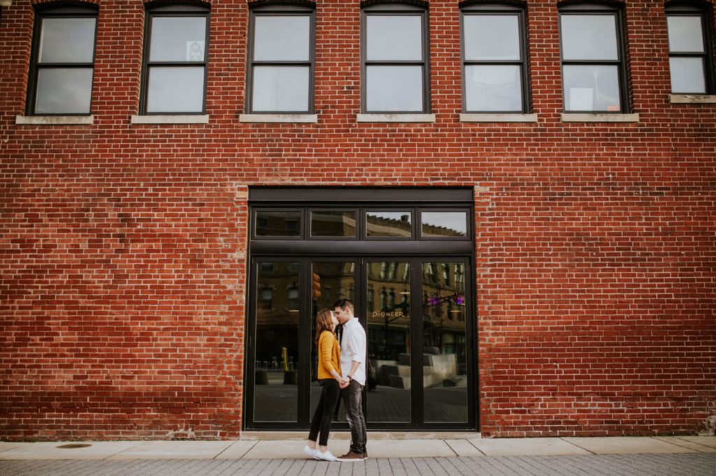 man and woman kiss in front of brick wall and large glass door Fountain Square Engagement