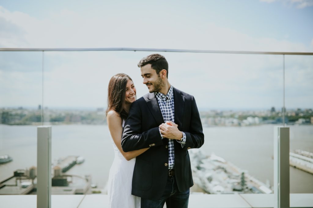 a young man in a jacket and a young woman in a white dress get New York rooftop engagement photos done in the midday sun