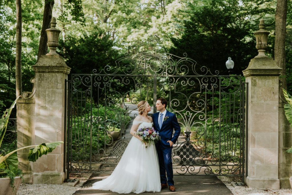 bride and groom stand in front of a metal gate in front of trees during their IMA wedding at newfields