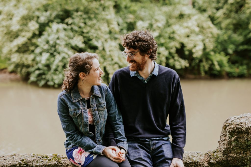man in sweater and woman in jean jacket sit on rough hewn stone wall during their Holcomb Gardens engagement shoot