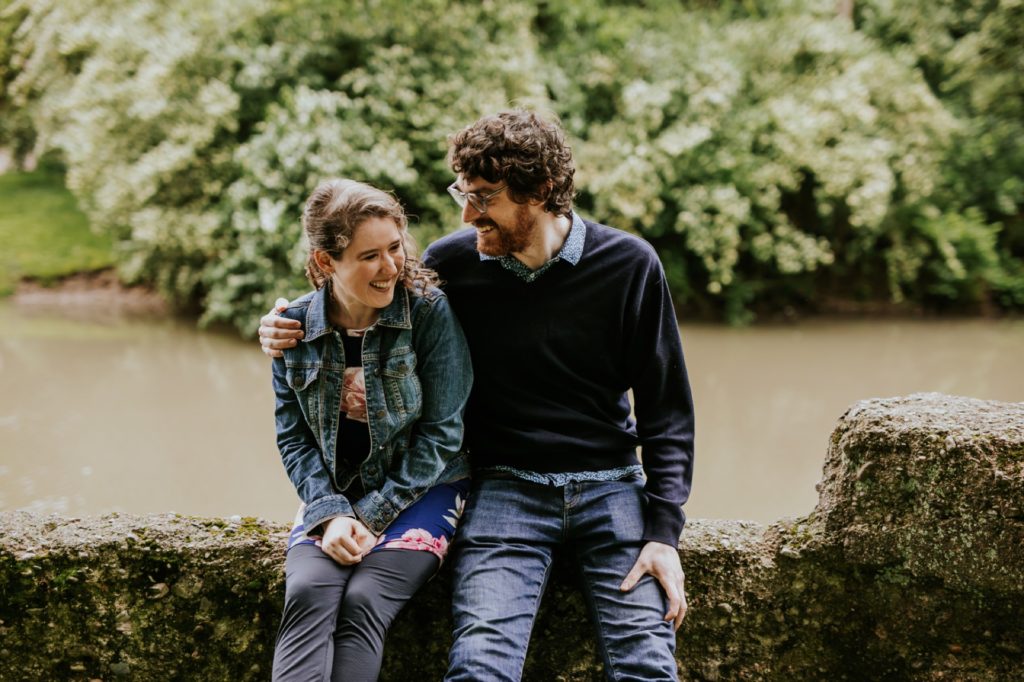 man in sweater hugs woman in jean jacket on rough stone wall at their Holcomb Gardens engagement shoot