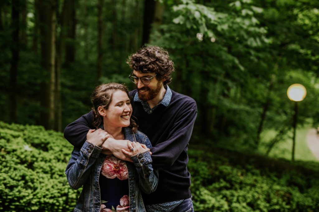 man with glasses hugs woman with floral print dress and jean jacket in woods at butler university