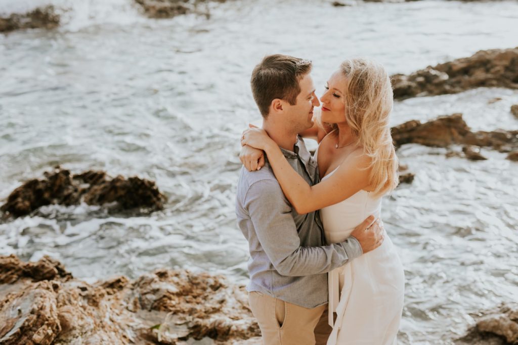 man and woman hug in front of swirling waters of the pacific ocean during their laguna beach engagement shoot