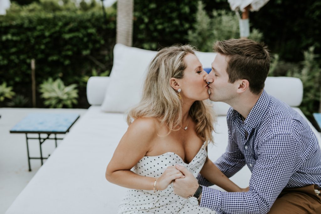 man and woman kiss each other on white chairs at hotel joaquin during their laguna beach engagement shoot