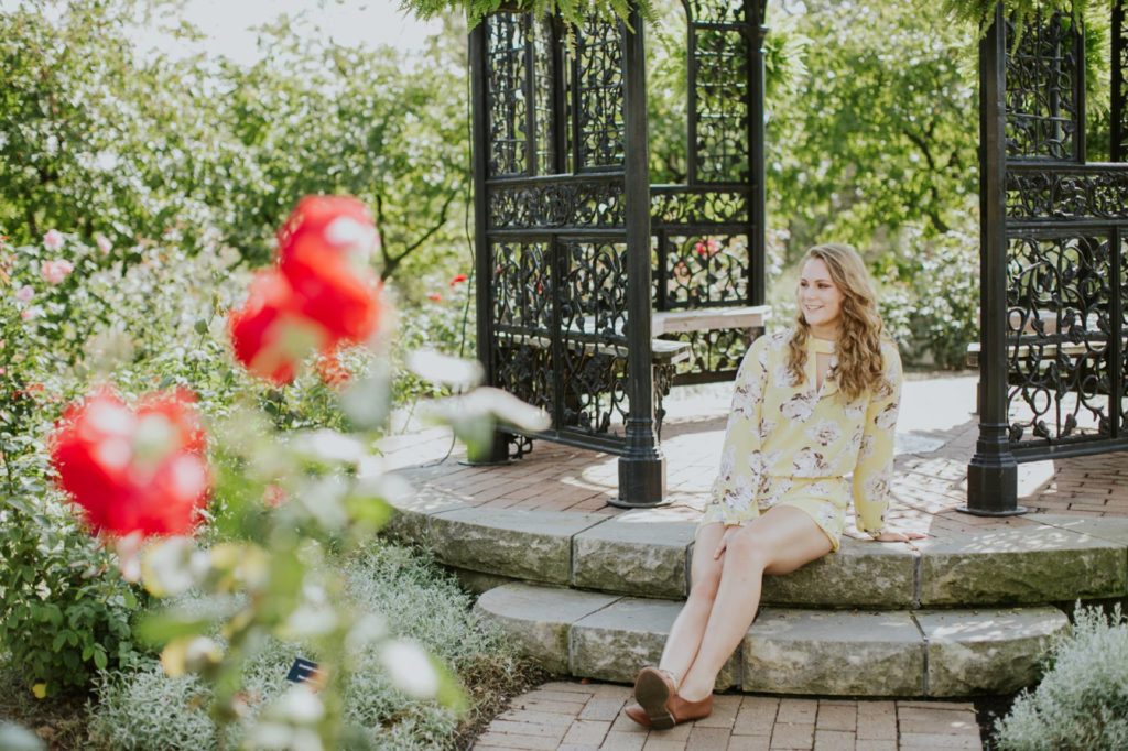 girl sits on steps of gazebo with red roses in foreground in her Muncie Indiana Senior Photography