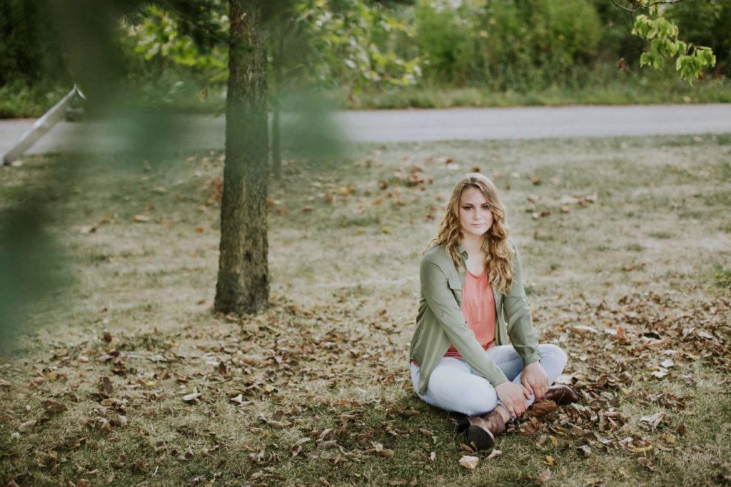 girl sits cross legged on grass surrounded by fallen leaves in this Noblesville Senior Photo Session