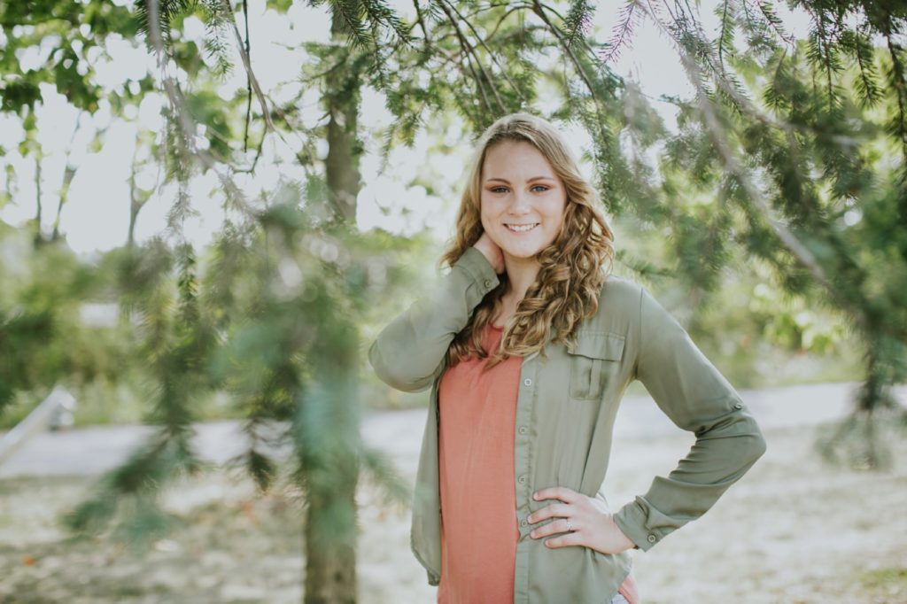 girl in orange shirt and green jacket stands under a tree with a hand on her hip Fishers Senior Photo Session