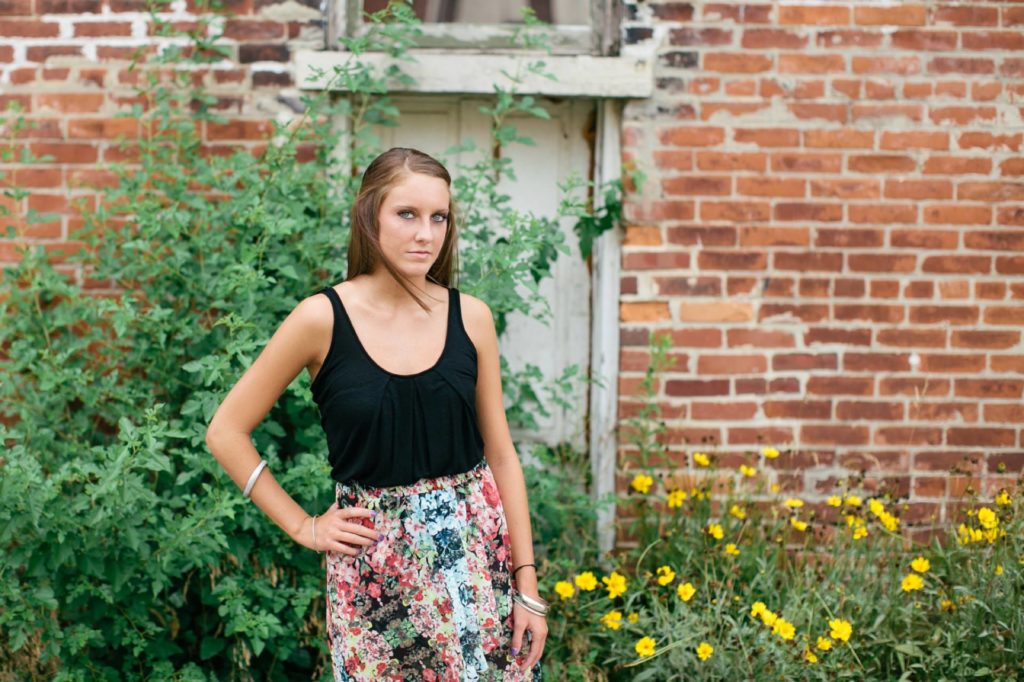 girl in black tank top with one hand on hip in front of brick wall with greenery in muncie senior photography