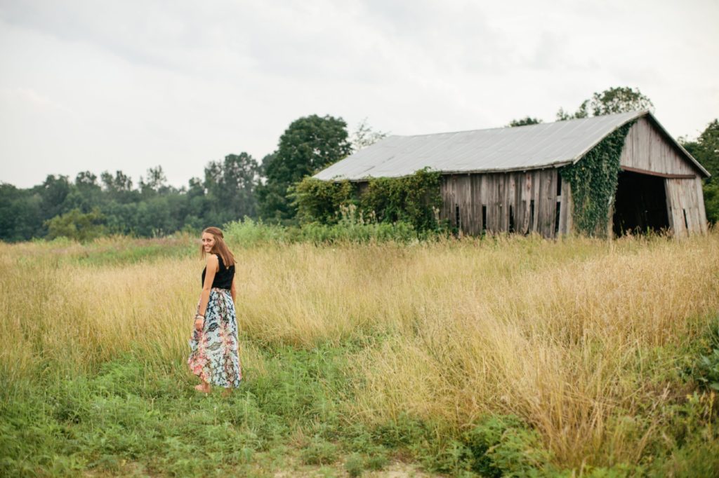 girl in black tank top and skirt walks through field in front of dilapidated barn in muncie senior photography