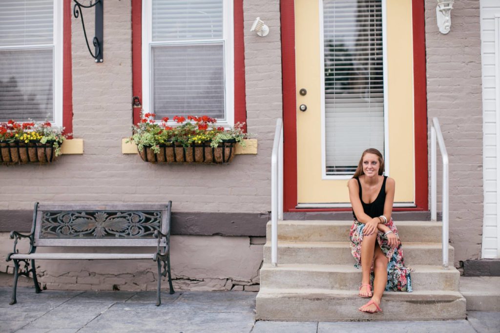 girl in black tank top sits on steps of house in front of yellow door with red framing in muncie senior photography