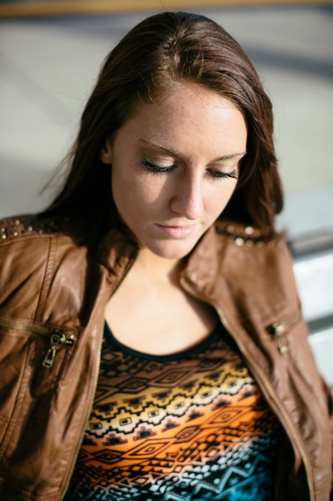 brunette girl in leather jacket looks down while sitting on bench during muncie senior pictures