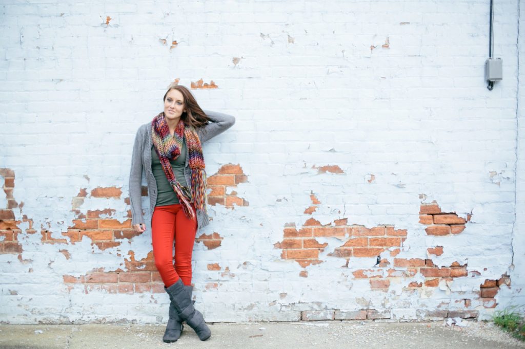girl in red pants and scarf leans against grungy brick wall with white paint in Noblesville portraits