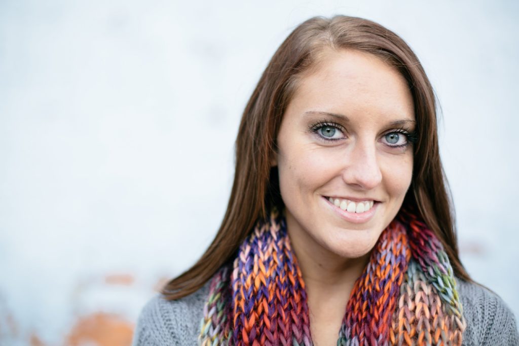 girl in scarf smiles in front of white brick wall in muncie senior pictures
