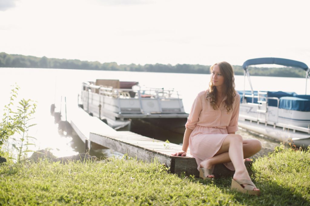 Girl in pink dress sits on dock at Prairie Creek Reservoir in Muncie senior portraits