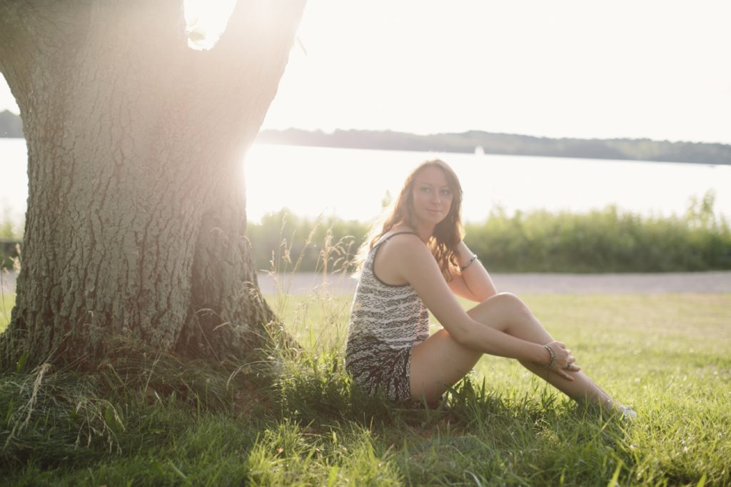 girl in tank top backlit by sun sits at base of tree near prairie creek reservoir in Muncie senior portraits