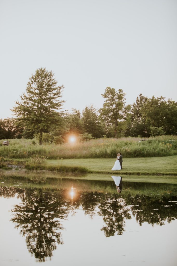 bride and groom stand on side of pond with sun setting behind them during their muncie wedding photography