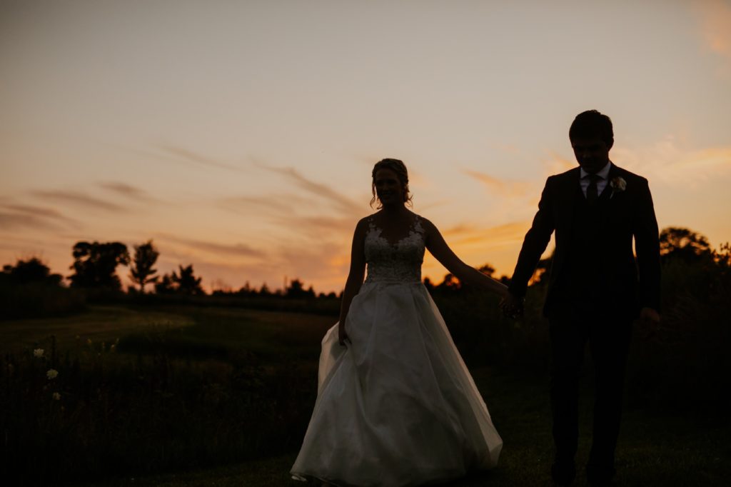 silhouette of bride and groom holding hands and walking away from sunset for their muncie wedding photography