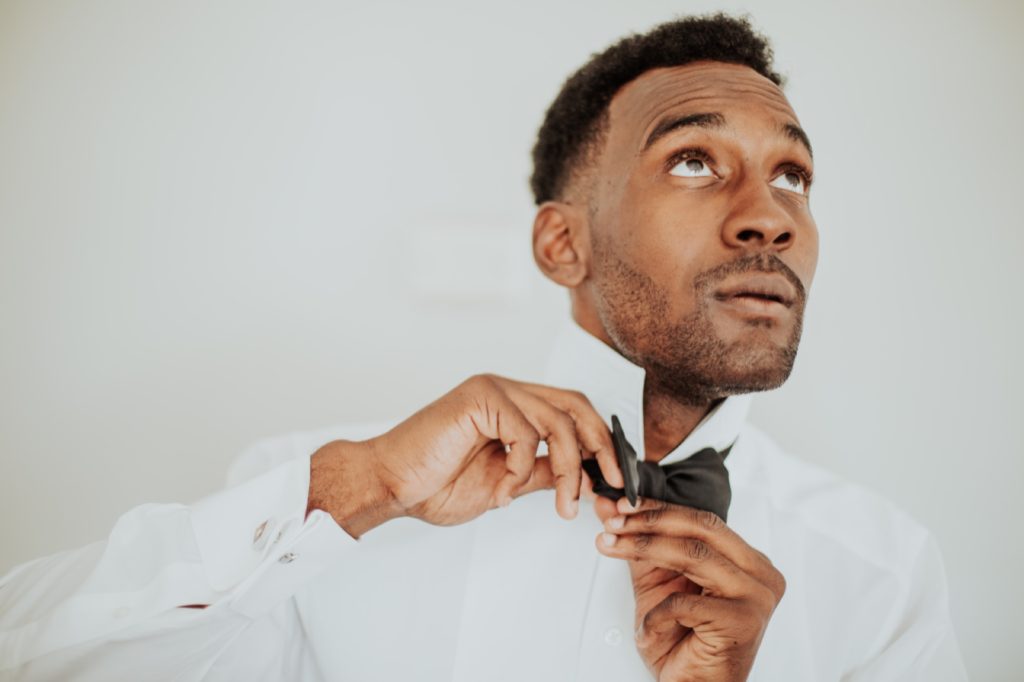 groom ties bow tie in front of white wall at airbnb in indianapolis