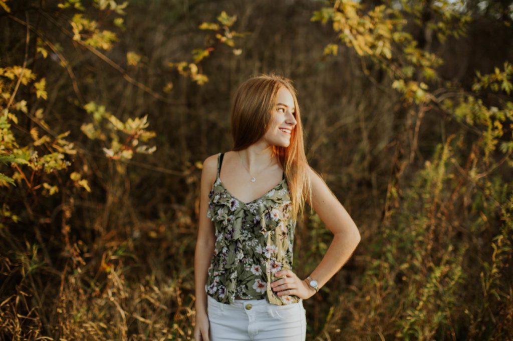 girl in floral tank top with hand on hip looks over her shoulder in the woods for Noblesville Senior Photos
