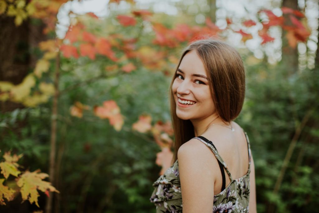 brunette girl in floral tank top smiles and looks over shoulder in front of orange leaves in Carmel portraits