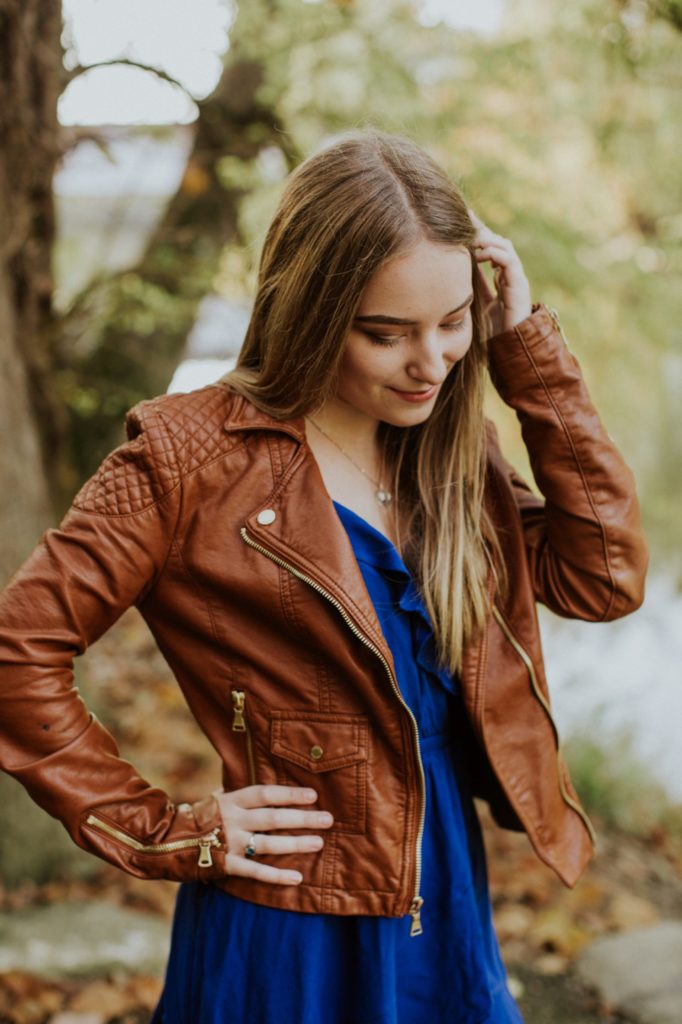 Girl in brown leather jacket in woods on waterfront for Noblesville Senior Portraits