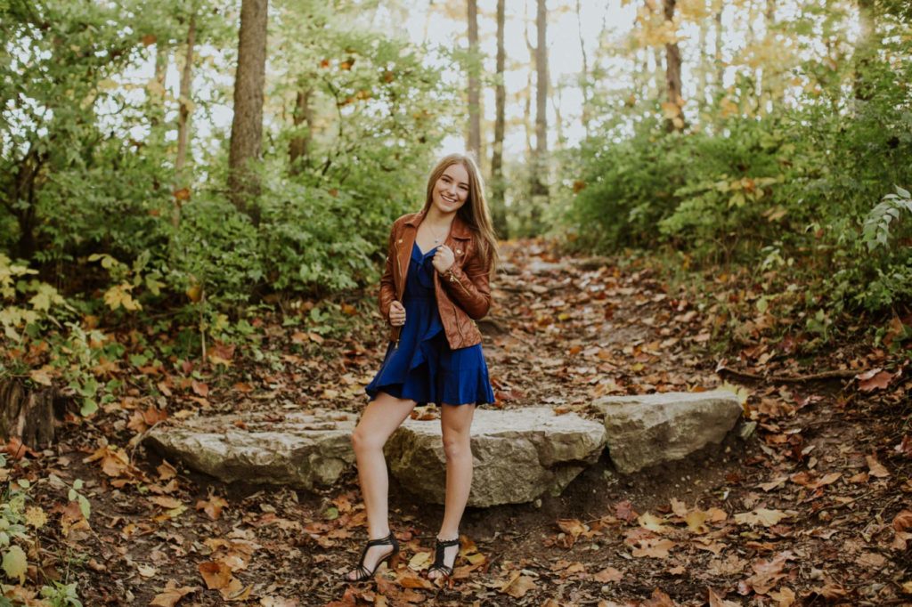 girl in brown leather jacket in dead leaves with light behind her for Noblesville Senior Portraits