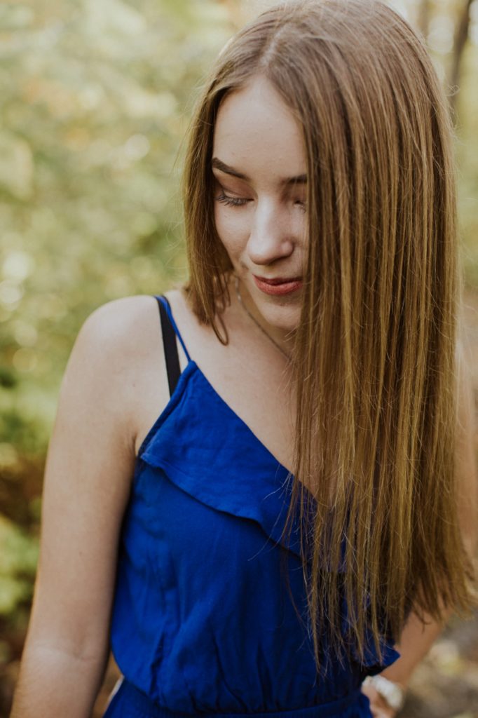 girl in blue dress with brown hair looking down in the woods for Noblesville Senior Portraits