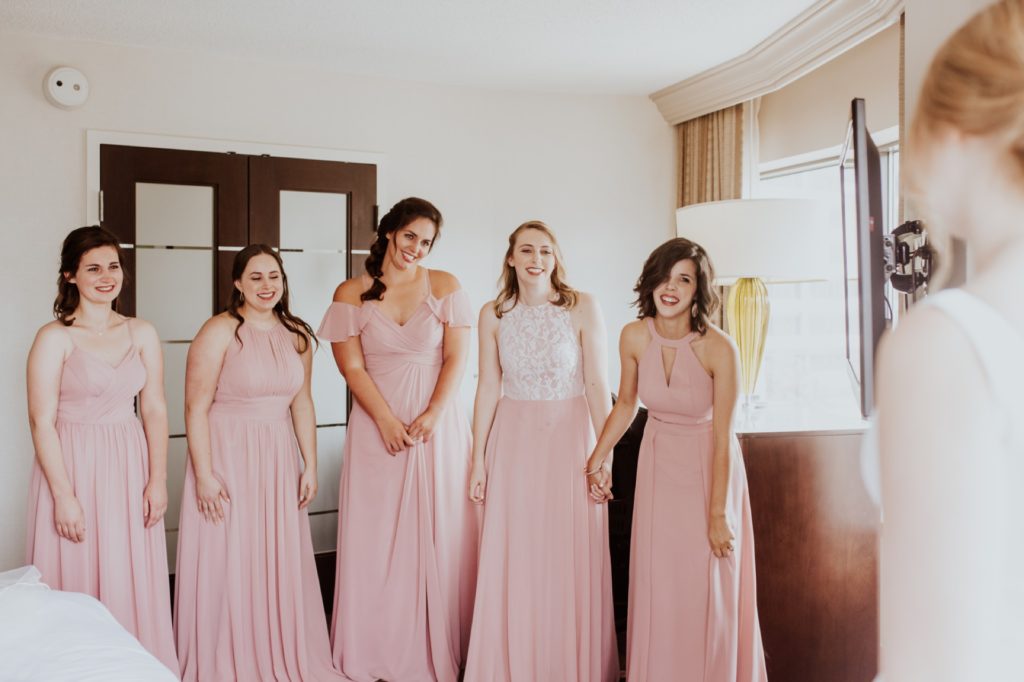 bridesmaids in pink dresses look lovingly at bride in hotel room