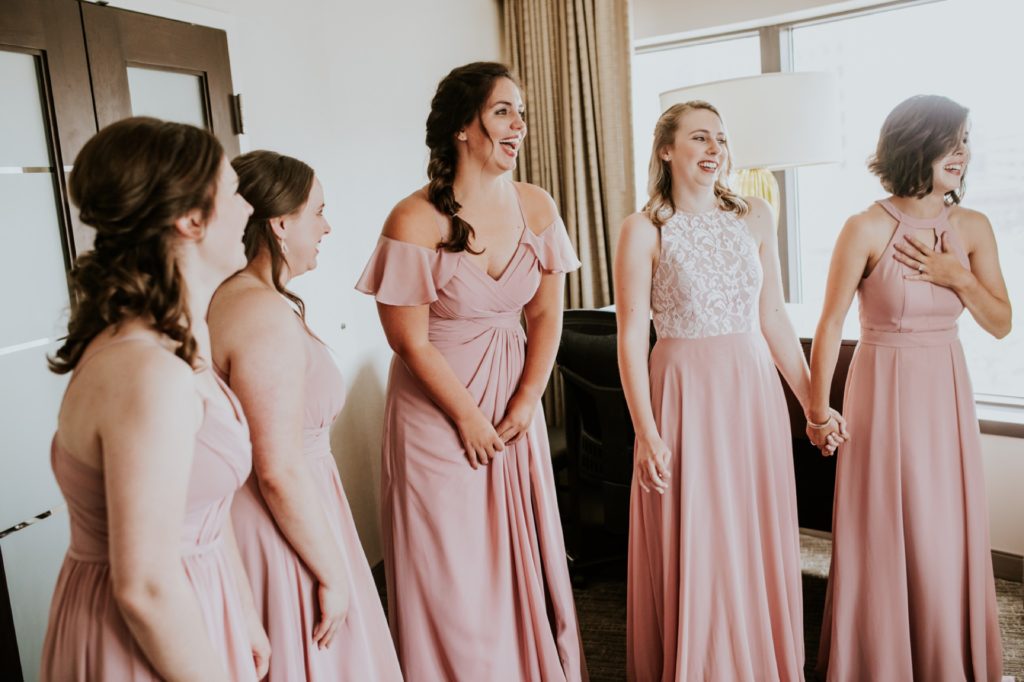 bridesmaids in pink dresses swoon as they look at the bride at a westin hotel room
