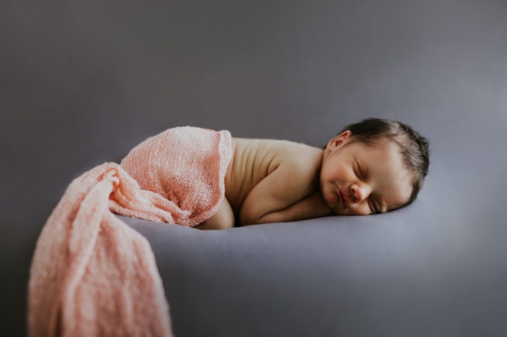New born baby girl in pink blanket sleeping on gray backdrop in carmel family portraits