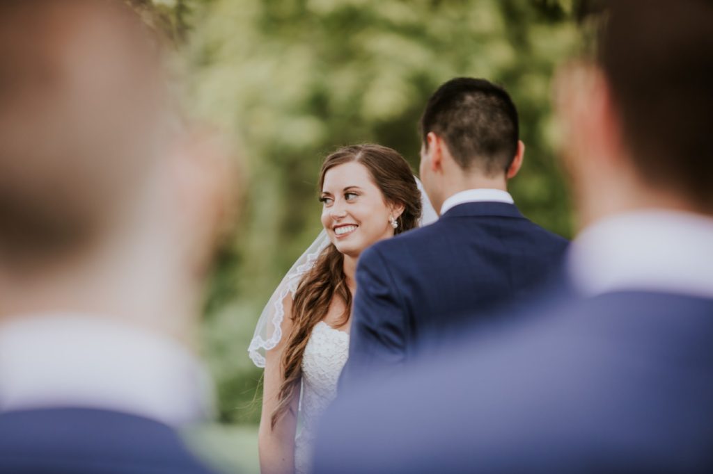 bride smiles as she is framed by groomsmen during ceremony at mustard seed gardens for Noblesville Wedding Photography
