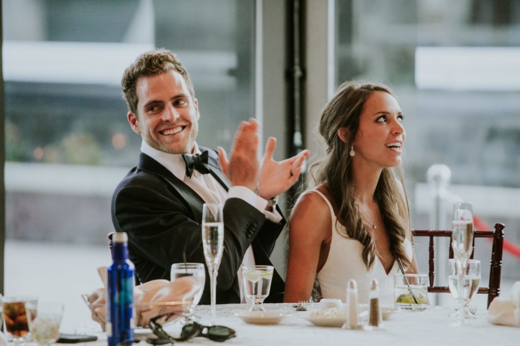 bride and groom sit in front of windows laughing and clapping during toasts for Indianapolis wedding photographer
