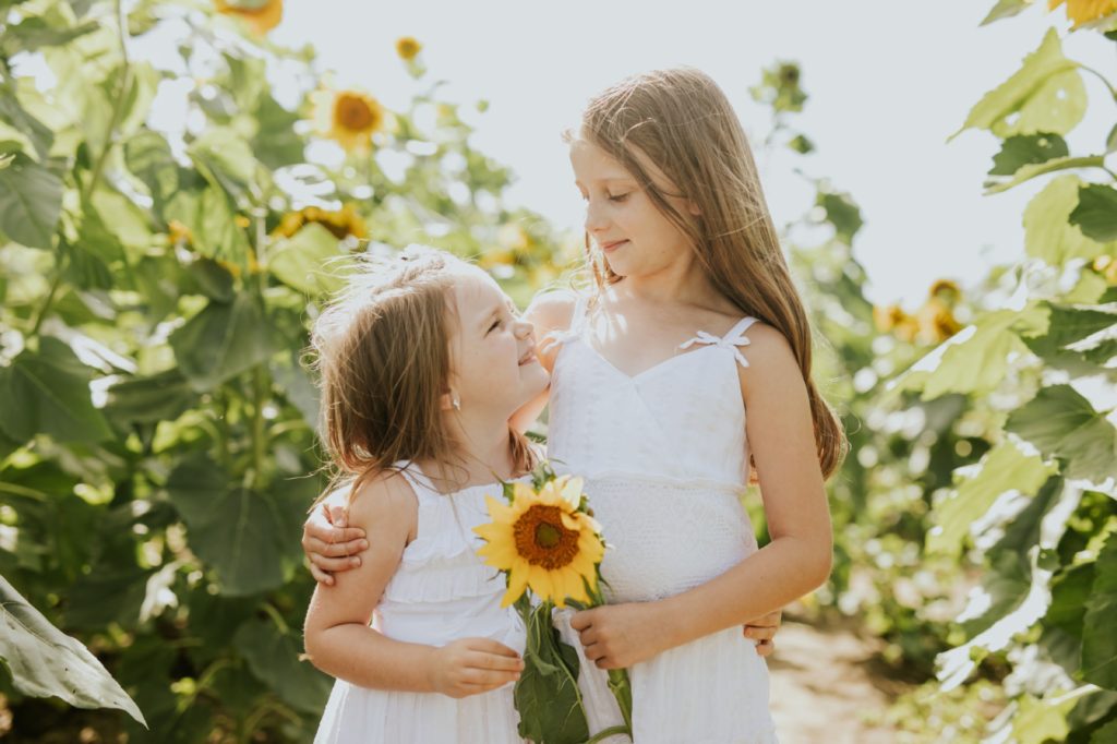 two girls in white dresses look at each other and smile while holding a sunflower at this Tuttle Orchards Family Photo session