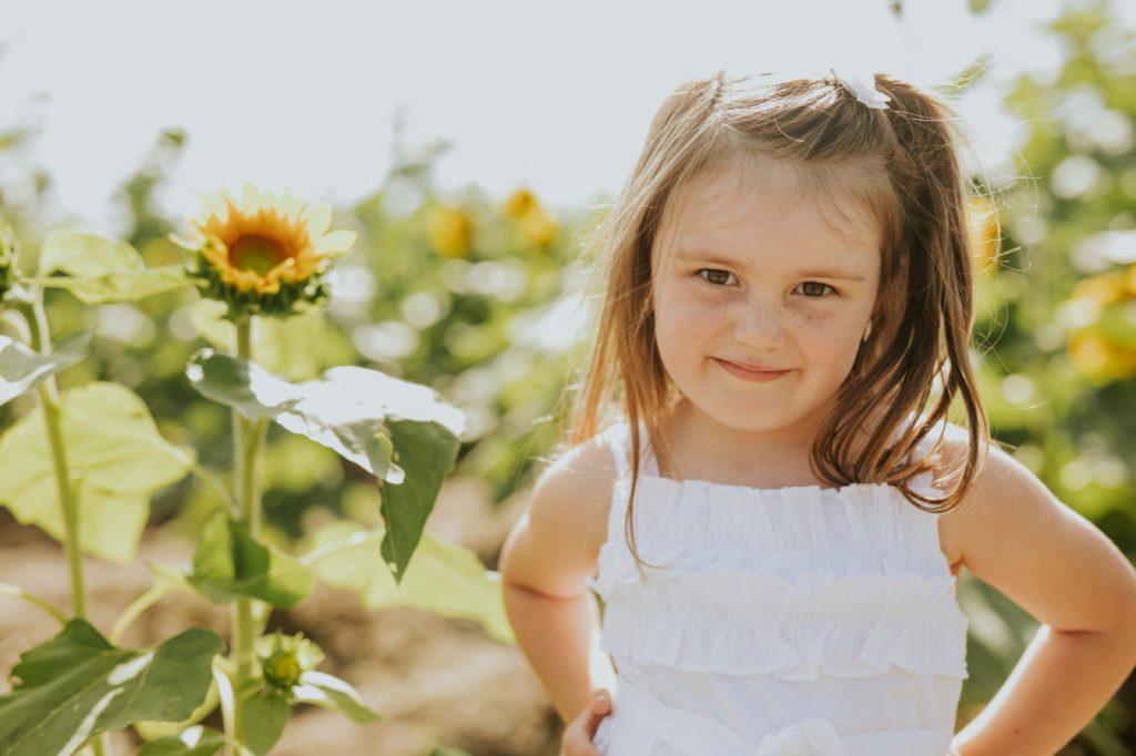 a young girl in a white dress stands with hands on hip in a sunflower field in these indianapolis family photos