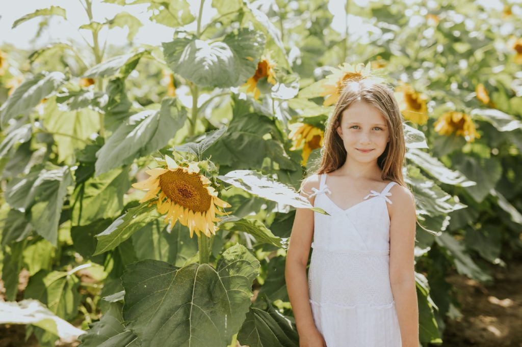 a young girl in a white dress stands next to a drooping sunflower in this Tuttle Orchards Family Photo session