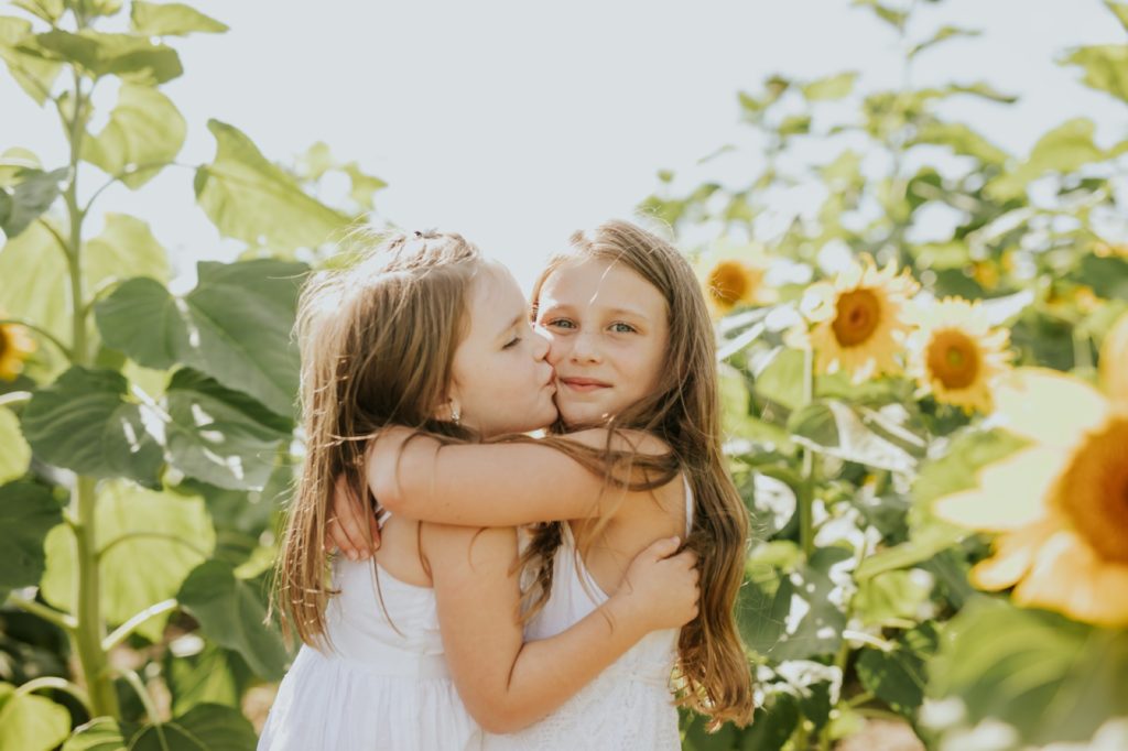 two young sisters in white dresses hug and kiss in a sunflower field in this Tuttle Orchards Family Photo session