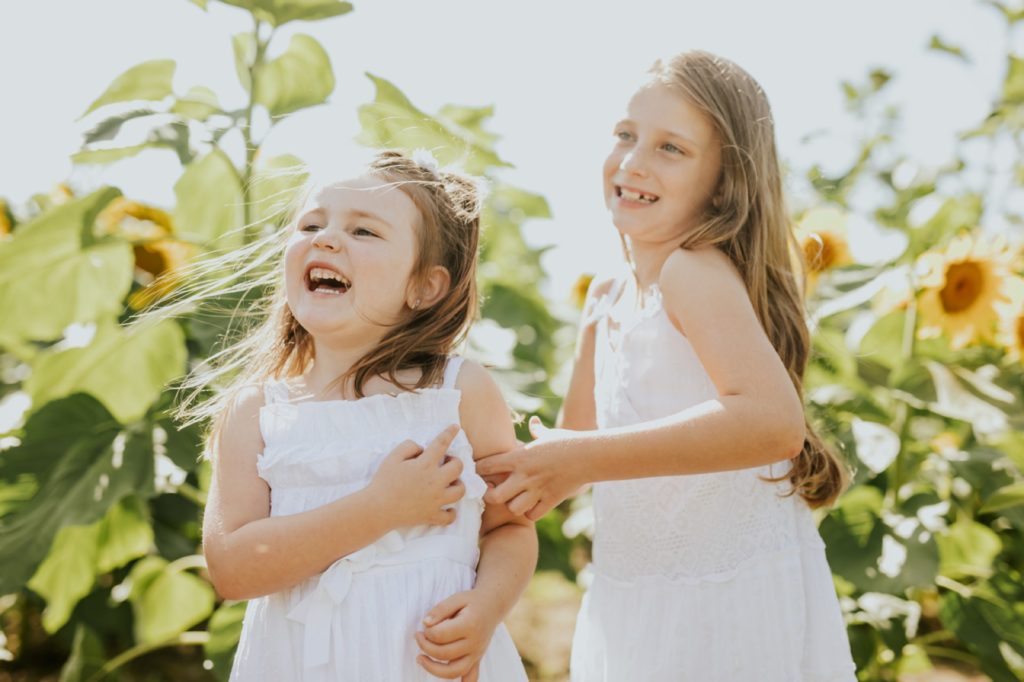 two sisters in white dresses laugh and run through a sunflower field in this noblesville family photo session