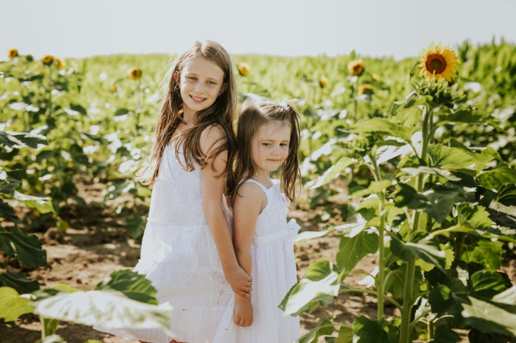 two girls stand back to back in a sunflower field holding hands in this carmel family photo session
