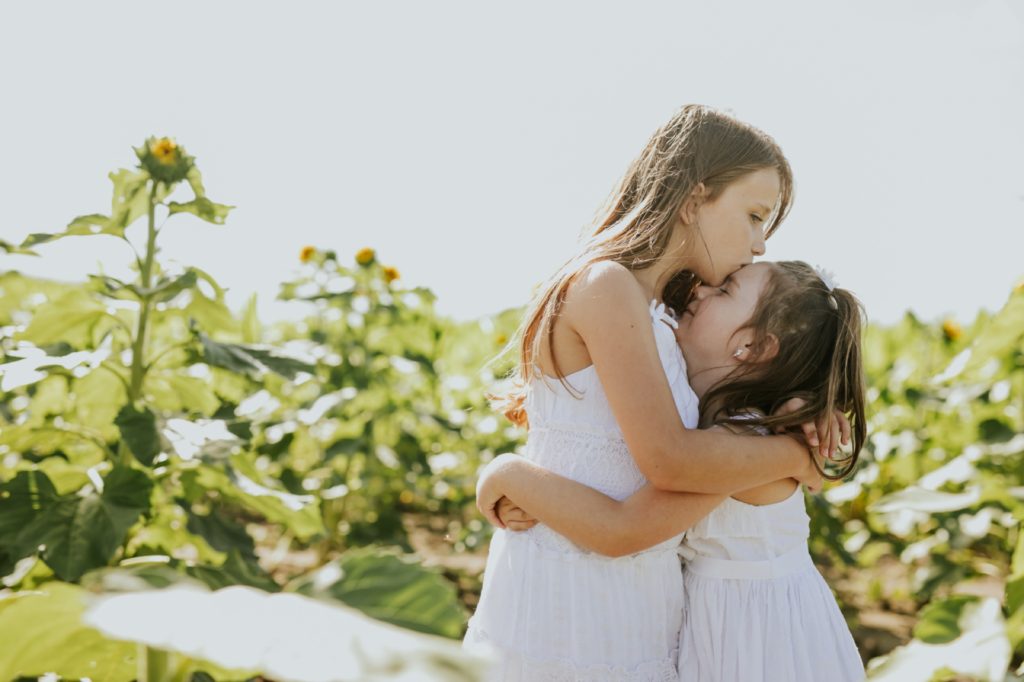 a young girl in a white dress kisses her sister on the forehead in a sunflower field in this zionsville family portraits