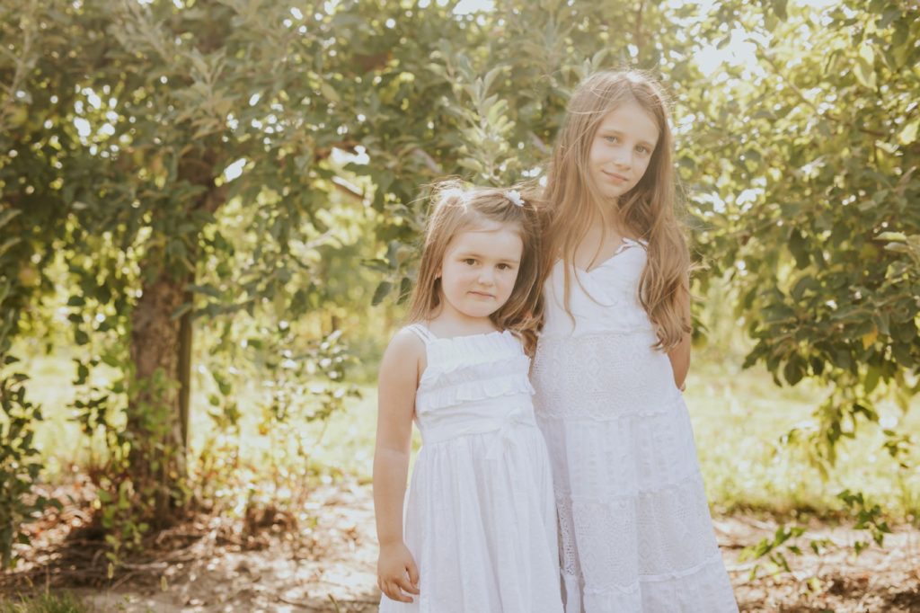 two girls in white dresses stand under an apple tree at sunset in this mccordsville Family Portraits