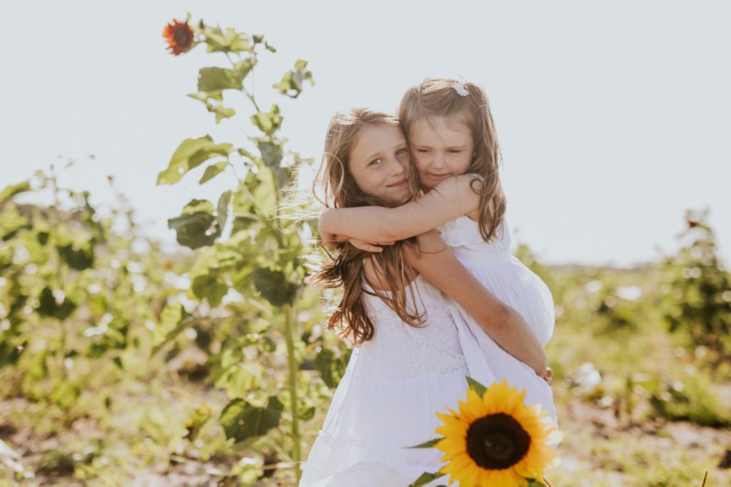 two girls in white dresses hug in a sunflower field in this mccordsville Family Photo session