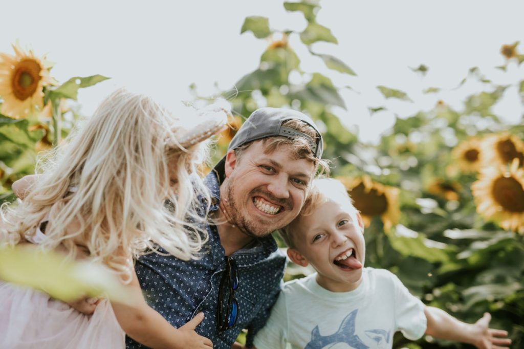 dad with backwards hat hugs two kids who are acting goofy in sunflower pictures at tuttle orchard