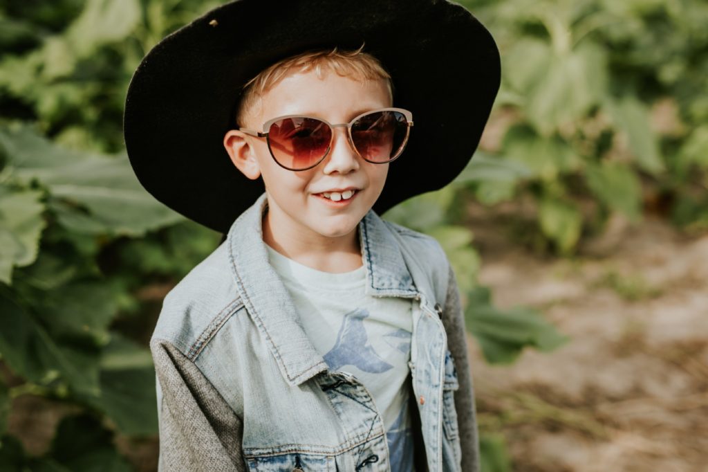 young blonde boy puts on his mother's denim jacket, floppy black hat and sunglasses in sunflower pictures at tuttle orchard
