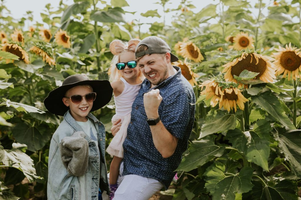 dad with backwards hat and scruffy facial hair poses with two kids in sunflower pictures at tuttle orchard