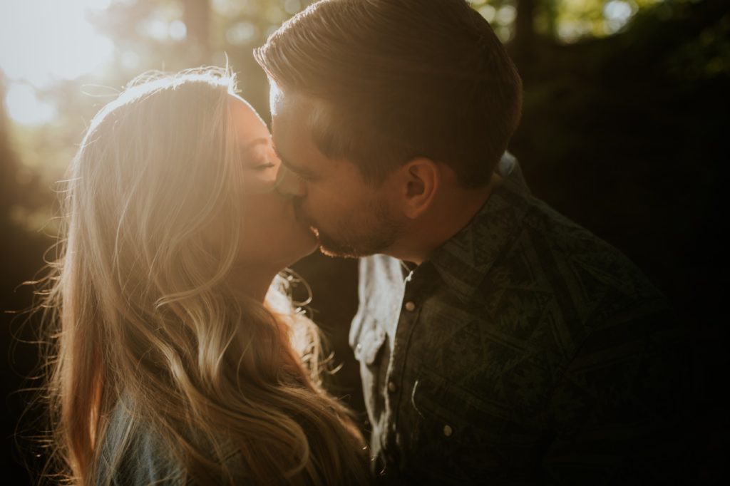 on ray of sunlight illuminates a man and a woman kissing in a dark woody area in this moody engagement portrait in indianapolis