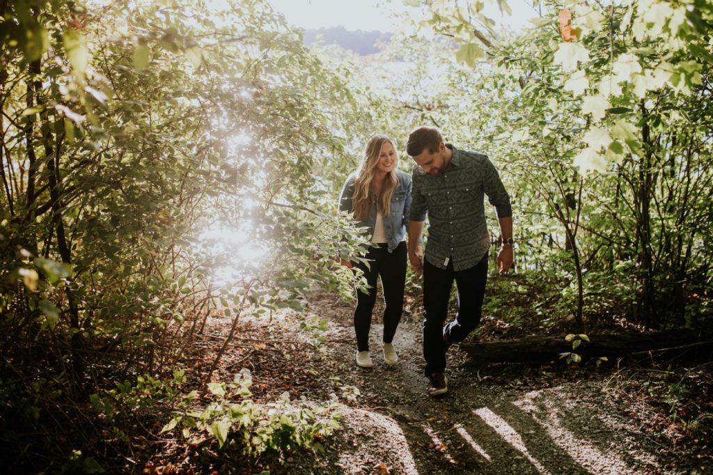 a man leads a woman by the hand as they walk down a path in the woods backlit by the sun